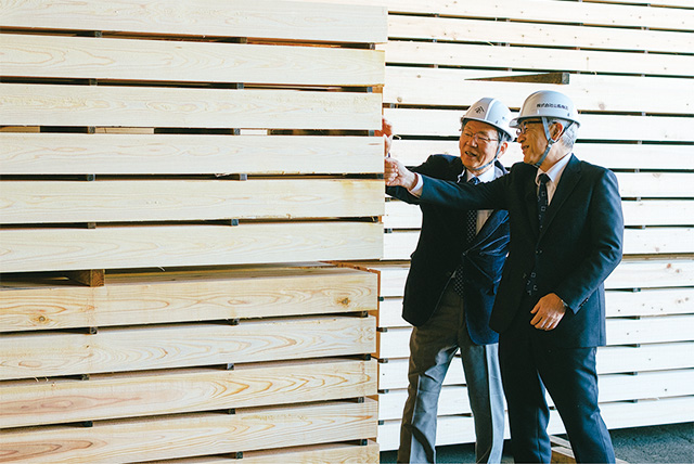 高く積み上げられた角材に手を触れながら話すヘルメットを被った二人の男性